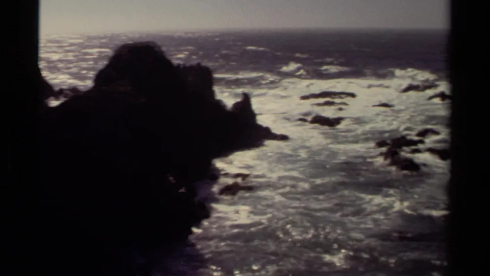 Faded 8mm coastal view from Mendocino with a large dark rock silhouette, foaming surf, and scattered offshore rocks.