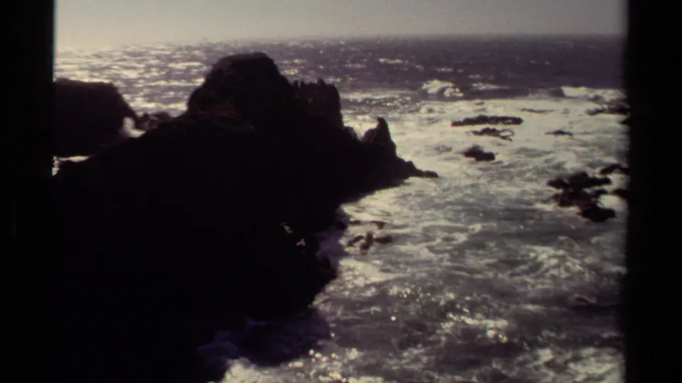 1980 8mm home-movie view of the Mendocino coast with dark rocks, bright surf, and hazy ocean light.