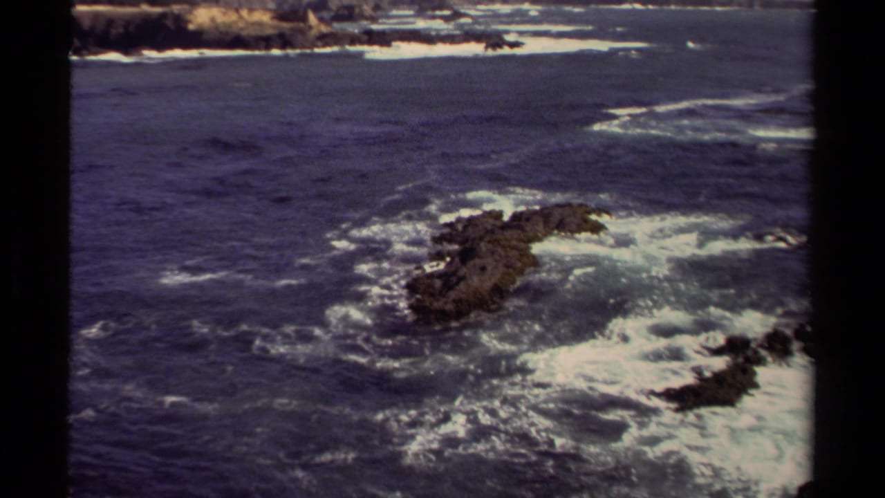 1980 8mm view of the Mendocino, California coastline, with surf curling around dark rocks under bright coastal light.