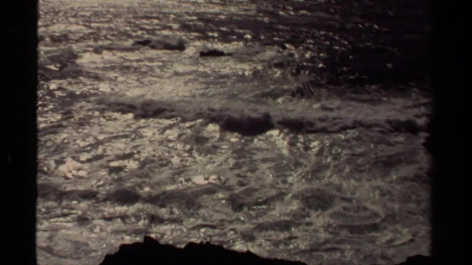 Vintage 8mm frame of churning Mendocino surf, low offshore rocks, and a dark foreground shoreline edge.