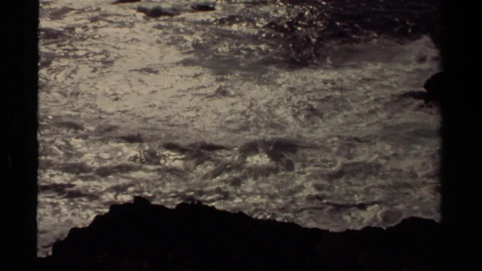 8mm frame of Mendocino waves and foam moving past a dark shoreline rock, with soft grain and subdued coastal light.