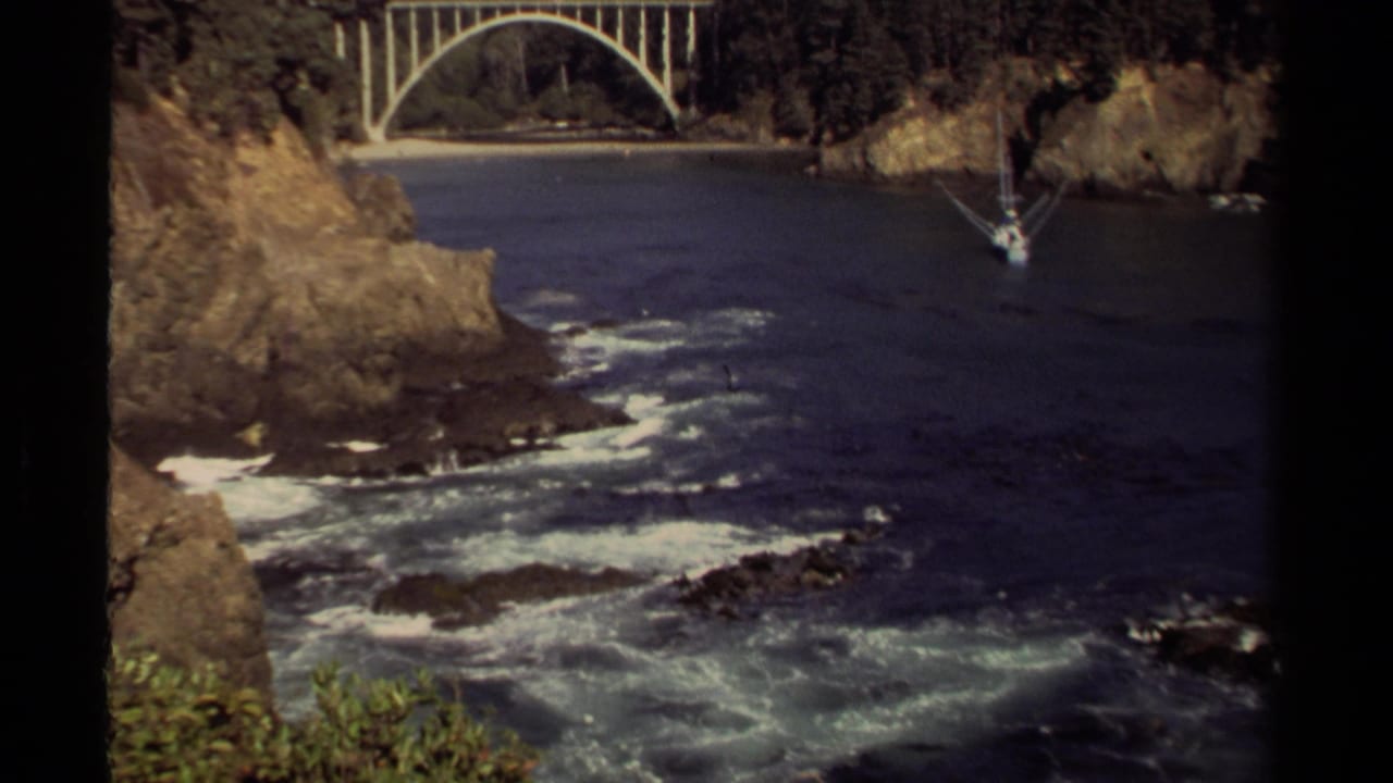Rocky coastal inlet with waves, an arched bridge, and a small sailboat near the cliffs.