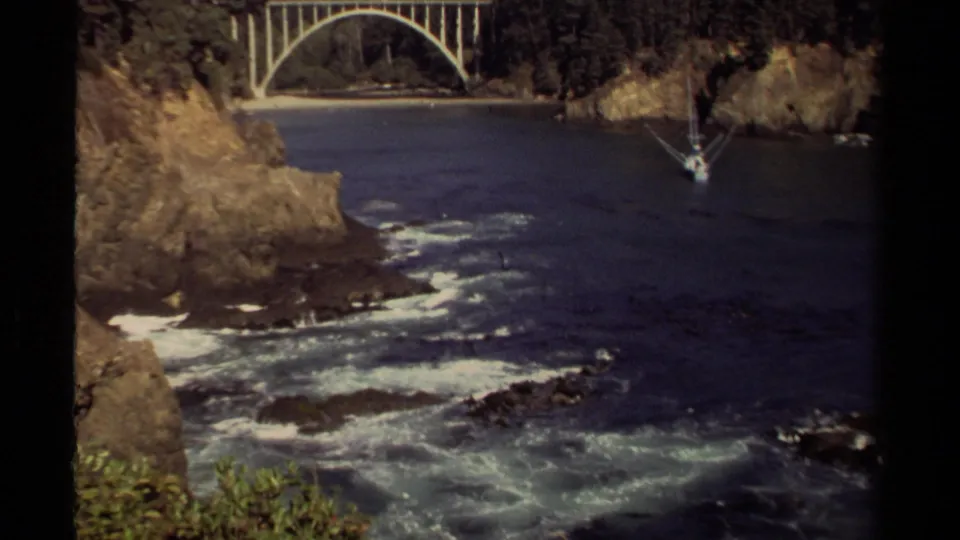 1980 8mm Mendocino coastal view showing white surf in a rocky inlet, an arched bridge, and a small sailboat near the far cliff.