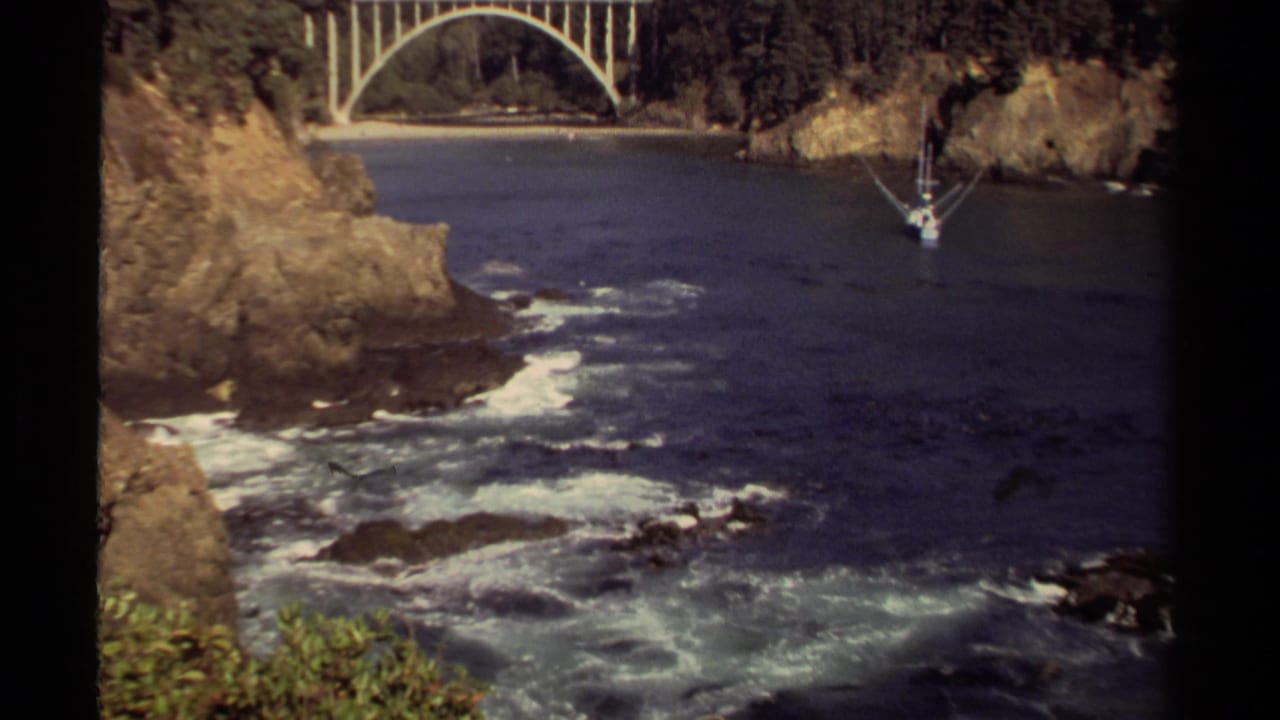 Mendocino coastal cove on 8mm film with surf moving through dark water, rocky cliff faces, bridge arch, and a small sailboat.