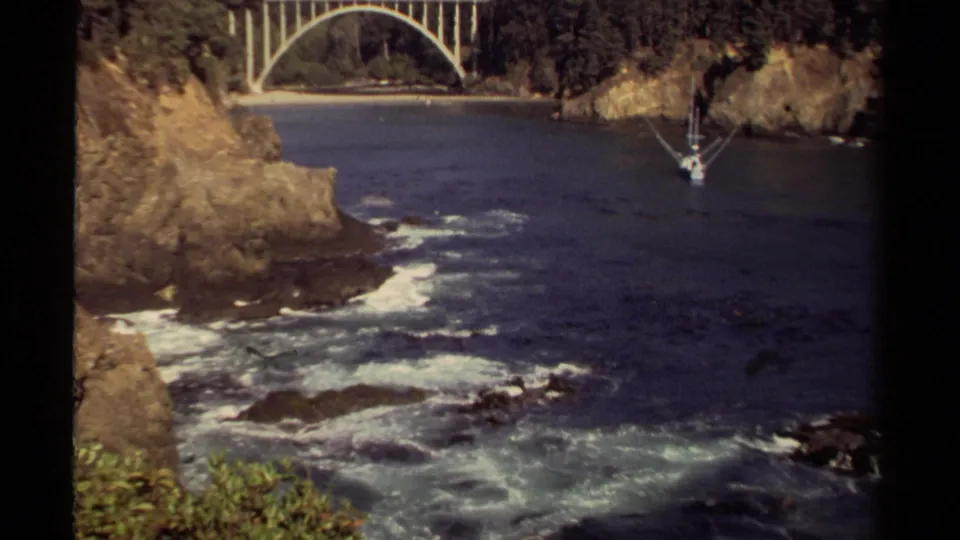 Mendocino coastal cove on 8mm film with surf moving through dark water, rocky cliff faces, bridge arch, and a small sailboat.