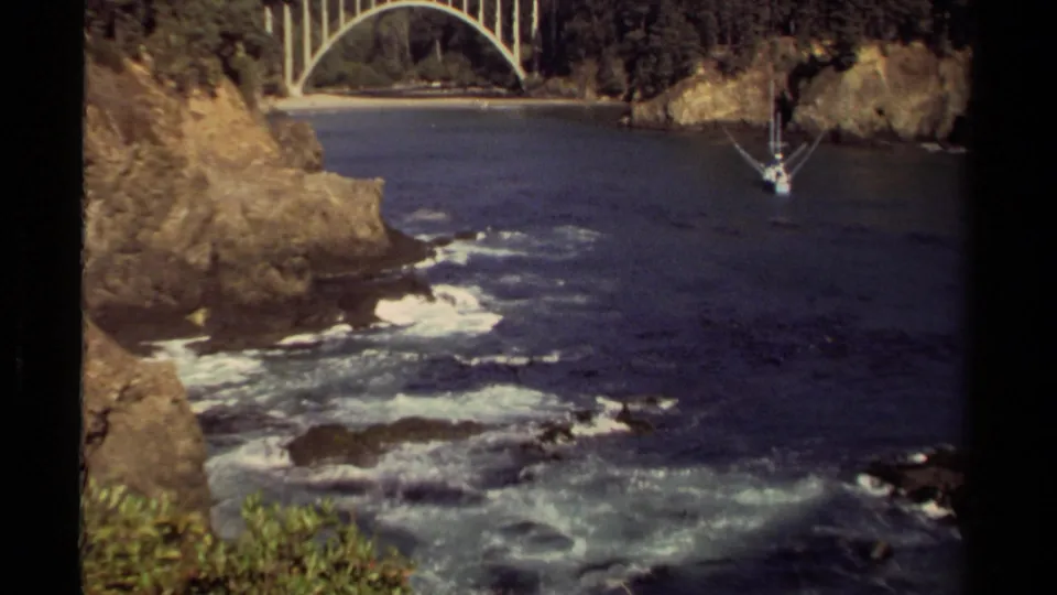 1980 Mendocino 8mm frame of a rocky inlet with foamy surf in the foreground, cliff edges, bridge structure, and a small sailboat beyond.