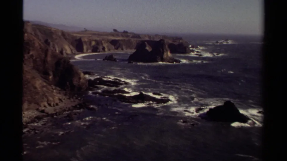 Wide 1980 8mm Mendocino coast view showing rugged cliffs, sea stacks, white surf lines, dark ocean water, and a hazy sky.