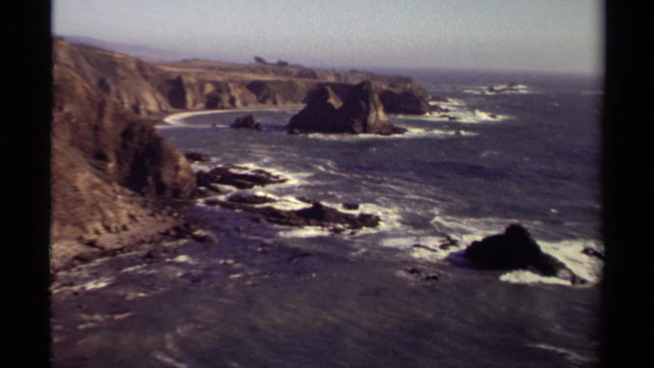 Mendocino coast seen from above, with rugged cliffs, offshore rocks, white wave lines, and open Pacific water in a soft 8mm frame.