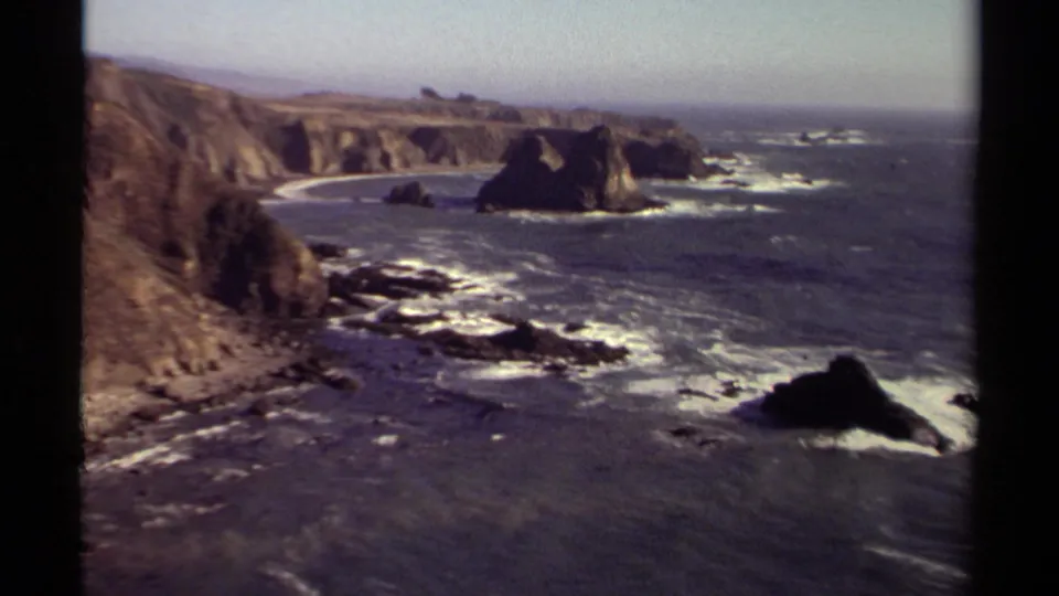1980 home-movie frame of the Mendocino shoreline, showing cliff faces, foamy surf around rocks, and a hazy ocean horizon.