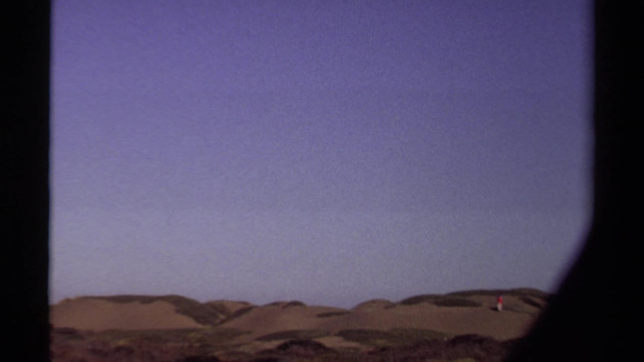 Distant person in red walking across sand dunes beneath a pale sky.