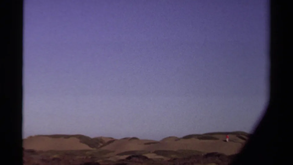 1980 Mendocino 8mm frame showing a distant person in red on rolling sand dunes under a broad pale-purple sky.