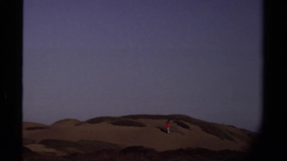 A zoomed 8mm view shows the lone walker in red and light pants moving across Mendocino sand dunes.