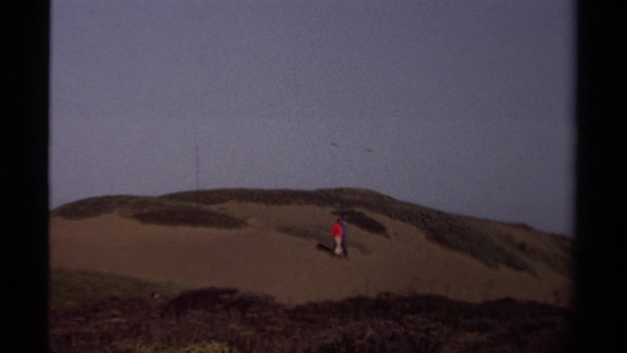 In supported 1980 Mendocino footage, two small figures cross coastal dunes with dark scrub and a dusky sky behind them.