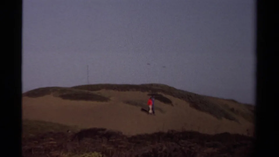 In supported 1980 Mendocino footage, two small figures cross coastal dunes with dark scrub and a dusky sky behind them.