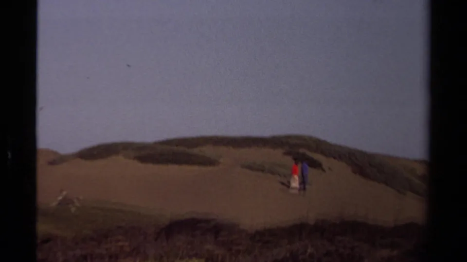 Two distant figures in colorful clothing appear on Mendocino coastal dunes, framed by scrub patches and a muted evening sky.