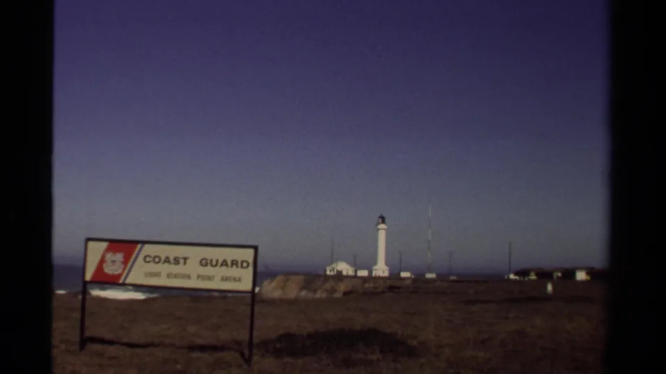 A Coast Guard sign appears in the foreground with a white lighthouse, low buildings, ocean, and cliffside terrain beyond in Mendocino, California.