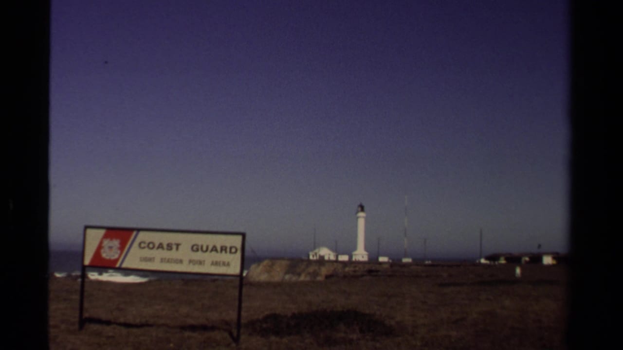 Coast Guard sign in front of a distant white lighthouse on a coastal bluff.