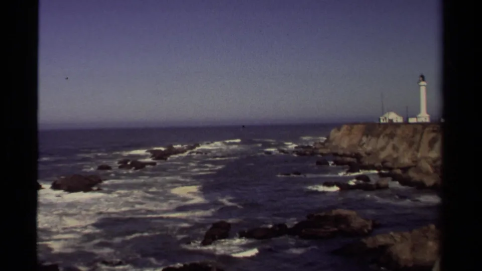 1980 8mm wide coastal view with Point Arena Lighthouse at right, Pacific waves below, and a clear horizon beyond.
