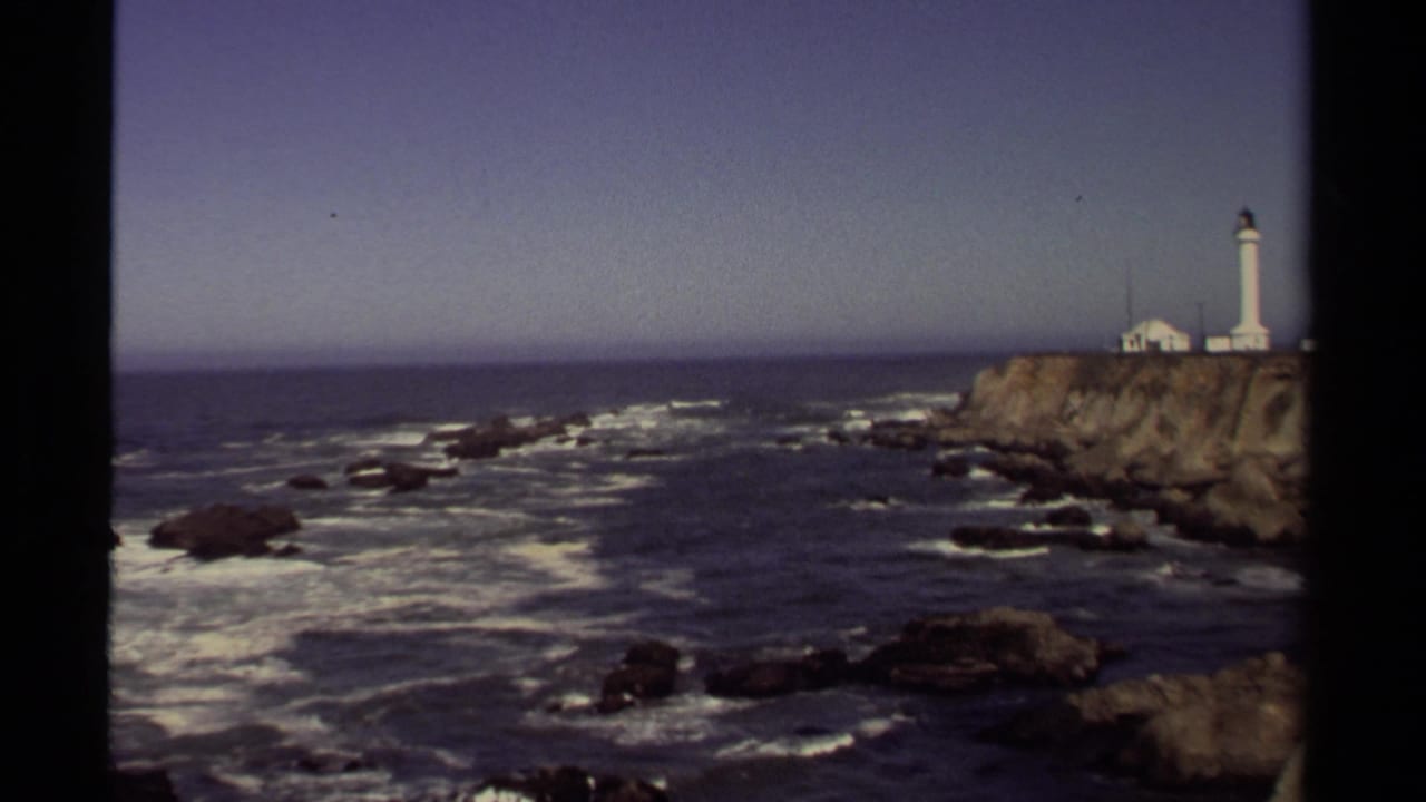 Wide 8mm frame of the Point Cabrillo Lighthouse complex on the Mendocino shoreline, seen across surf and scattered rocks.