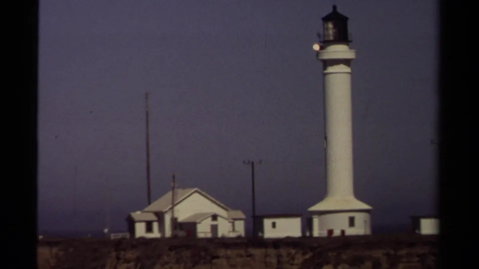 Close 8mm frame of the Mendocino coast lighthouse complex, with the white tower rising beside low buildings on the bluff.