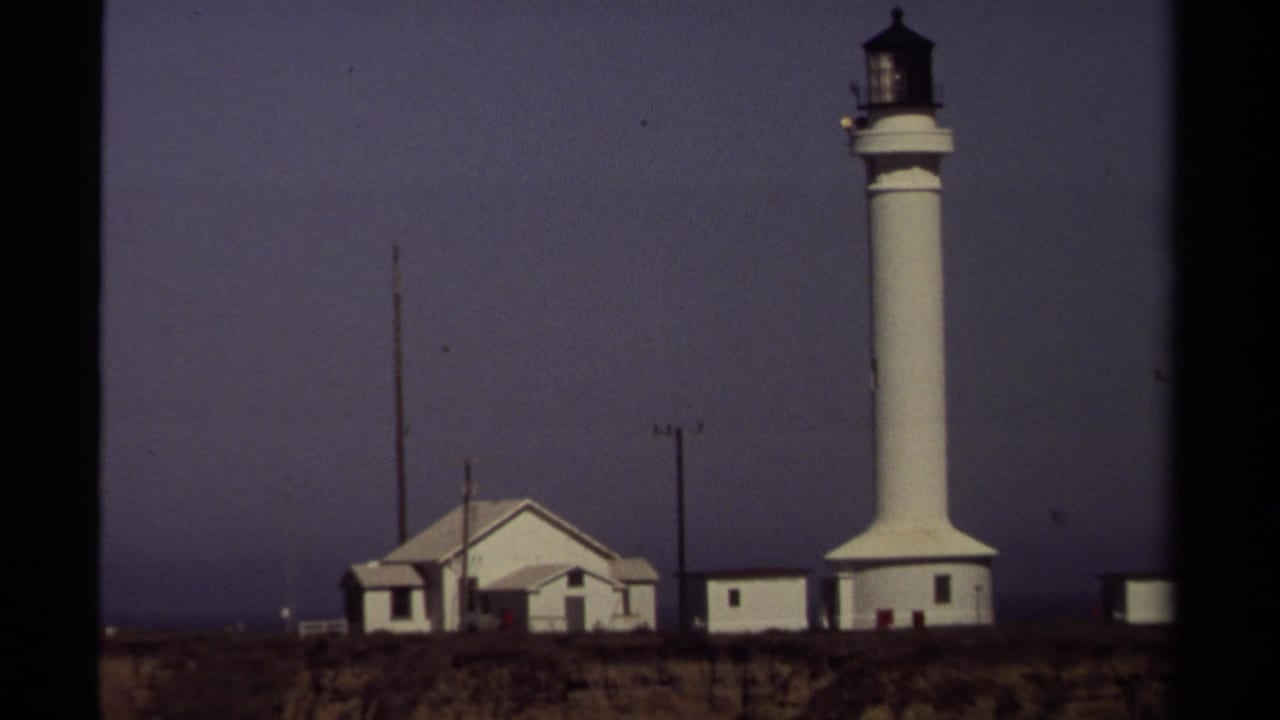 White lighthouse and nearby buildings on a coastal cliff under a dark sky.