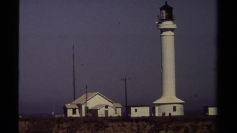 1980 8mm view of a Mendocino, California lighthouse and adjacent buildings seen from across a grassy cliff under an overcast sky.