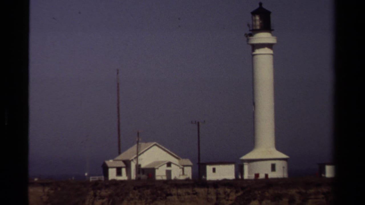 The Mendocino coastal lighthouse complex appears in a steady 8mm frame, with the tower, low buildings, poles, and dark horizon visible.