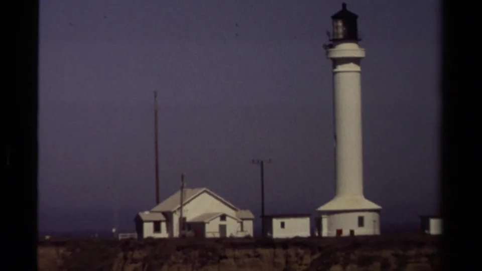 The Mendocino coastal lighthouse complex appears in a steady 8mm frame, with the tower, low buildings, poles, and dark horizon visible.