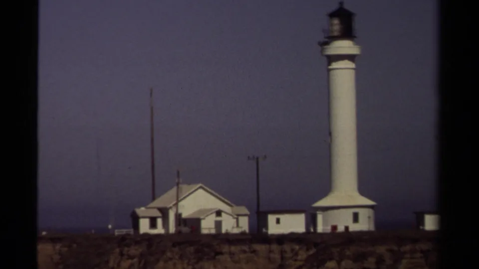 A closer 1980 8mm frame shows the white Mendocino lighthouse tower rising beside low buildings on the coastal cliff.
