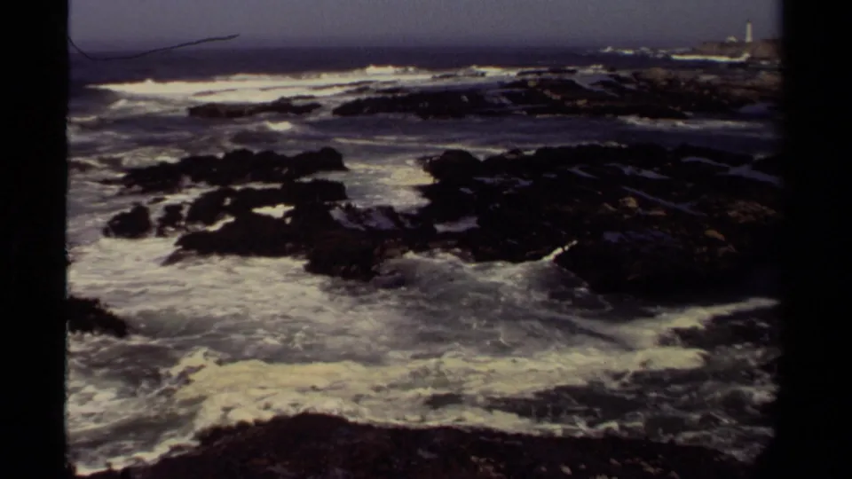 Foamy Pacific surf breaks across dark rocks on the Mendocino coast, with the lighthouse faintly visible in the far distance.