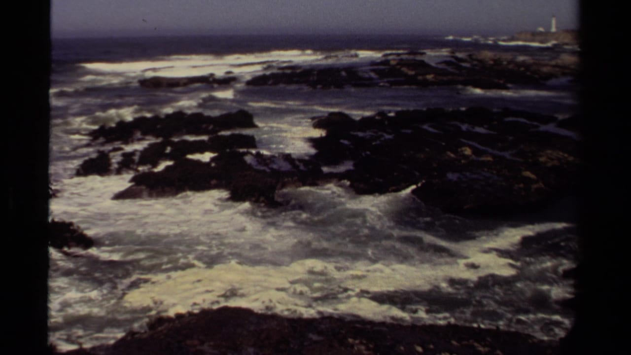 Large waves break over dark rocks with a distant headland and tower on the horizon.
