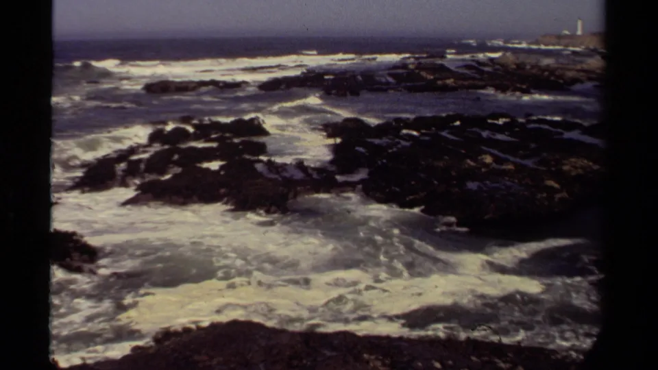 1980 Mendocino 8mm view of waves surging around low dark rocks, with a hazy Pacific horizon and distant coastal point in the background.