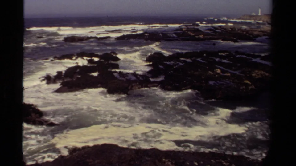 1980 Mendocino, California frame of rolling surf crossing black shoreline rocks, with a distant headland and lighthouse-like structure near the right edge.