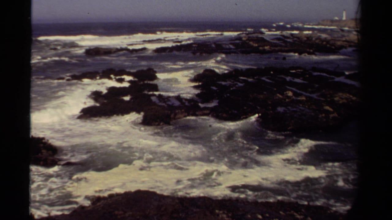 1980 Mendocino coastal home-movie frame showing waves over dark rocks with a small distant lighthouse near the horizon.