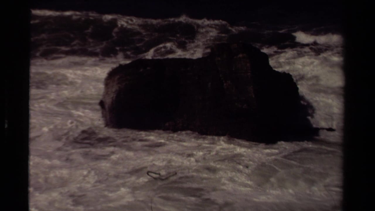 Large dark rock surrounded by turbulent white water and breaking waves.
