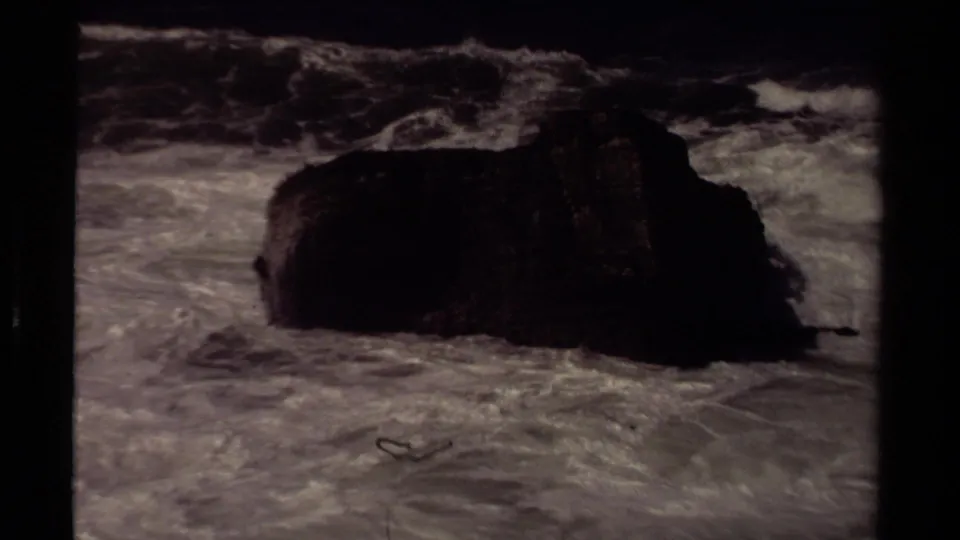 1980 Mendocino coast frame with a dark offshore rock surrounded by churning surf and muted film color.