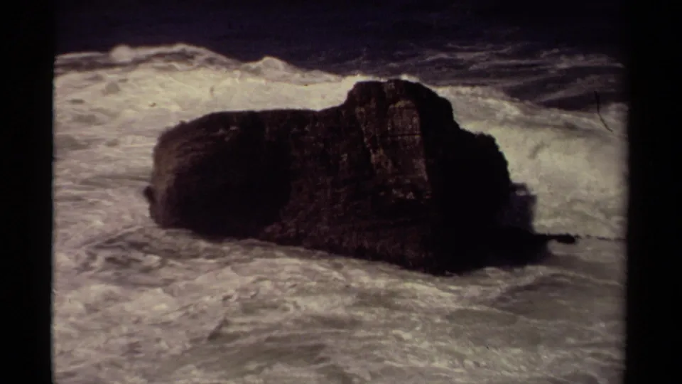 Vintage 8mm Mendocino shoreline frame showing foamy surf wrapping around a dark jagged rock.
