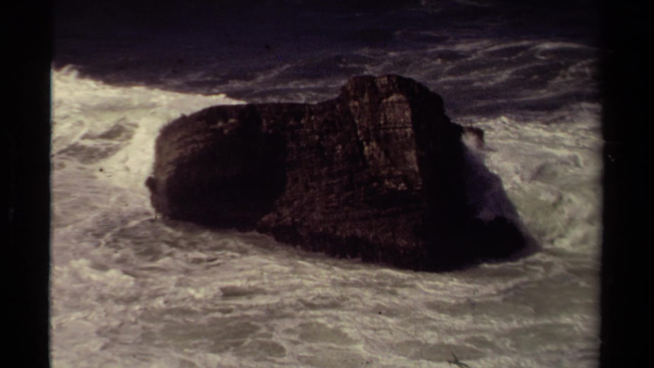 Dark offshore rock surrounded by white surf and churning ocean water.