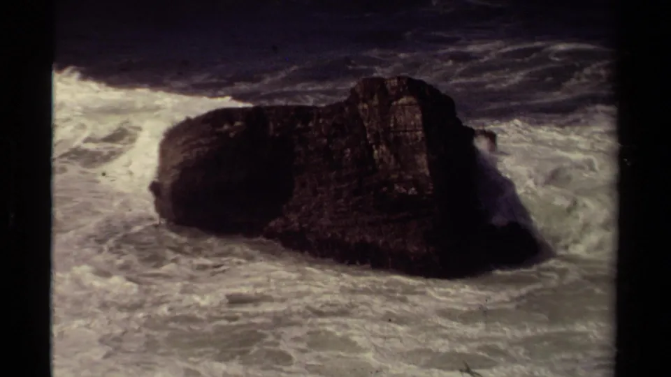 1980 Mendocino 8mm frame showing a dark rock formation in the ocean as white waves break around it.