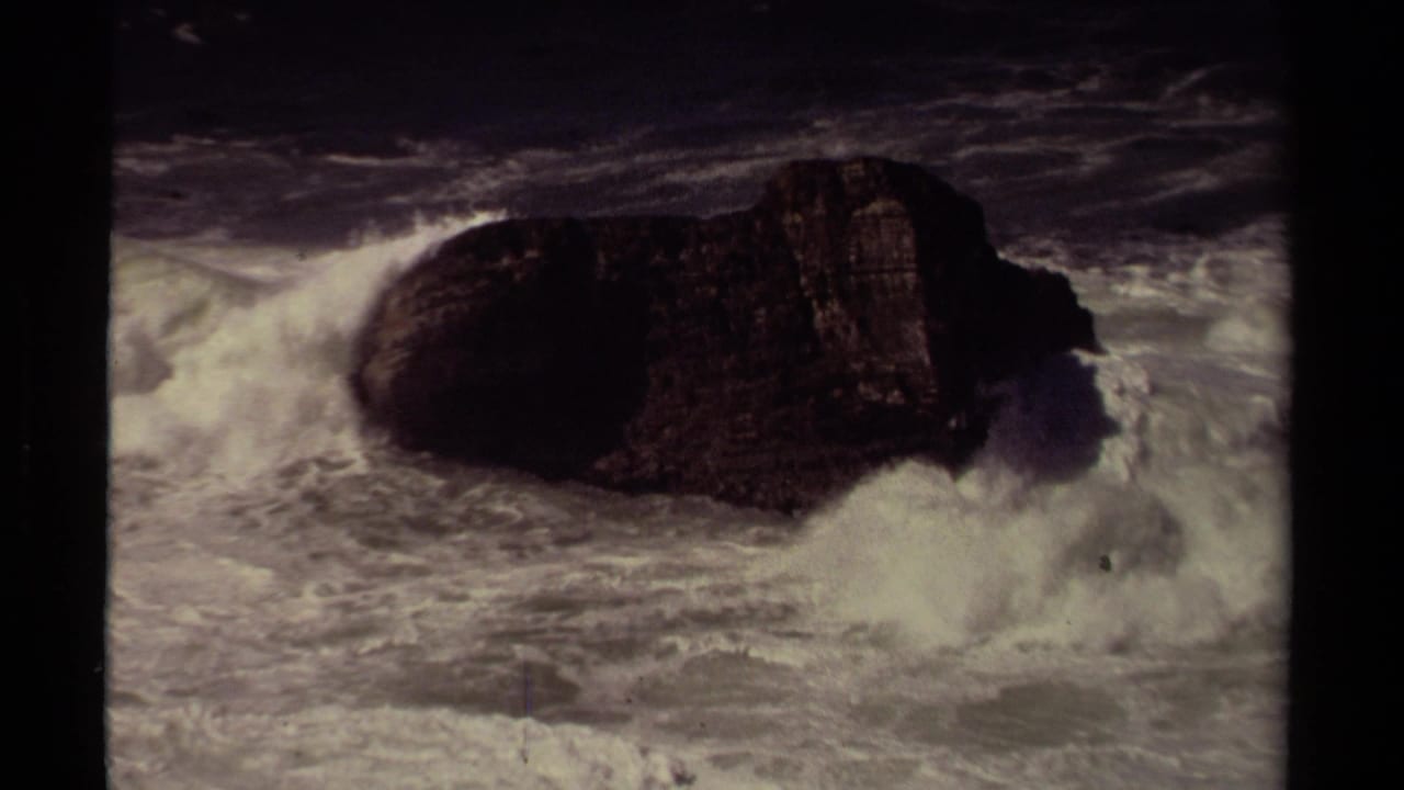1980 Mendocino 8mm frame with surf slamming into a dark offshore rock and throwing white spray upward.