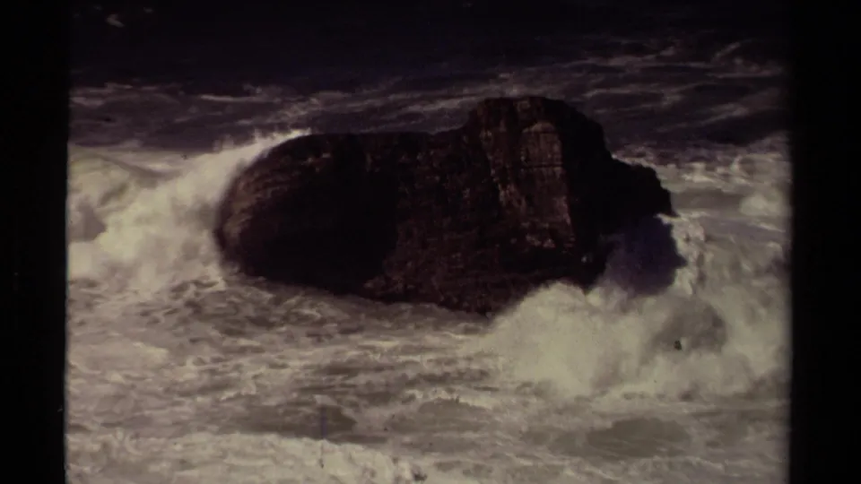 1980 Mendocino 8mm frame with surf slamming into a dark offshore rock and throwing white spray upward.