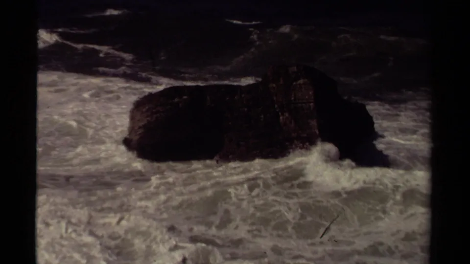 1980 Mendocino 8mm frame showing the rock partly shadowed while pale surf churns around its base.