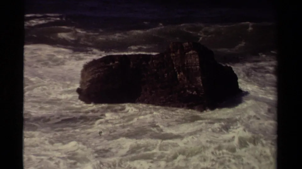 1980 Mendocino 8mm frame holding on the offshore rock as foamy Pacific water spreads across the foreground.