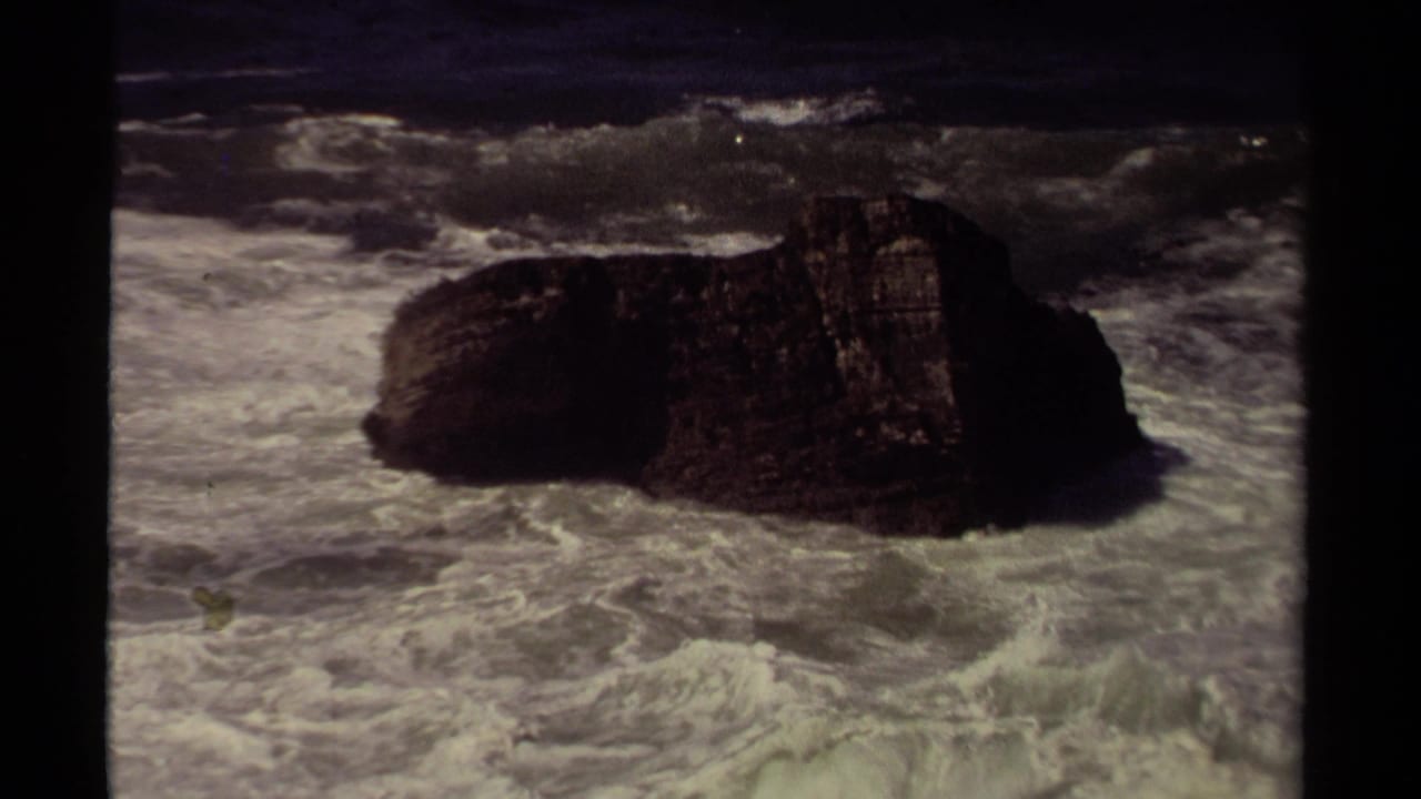 1980 8mm home-movie frame of a dark rock formation near the Mendocino coast with whitewash moving around it.