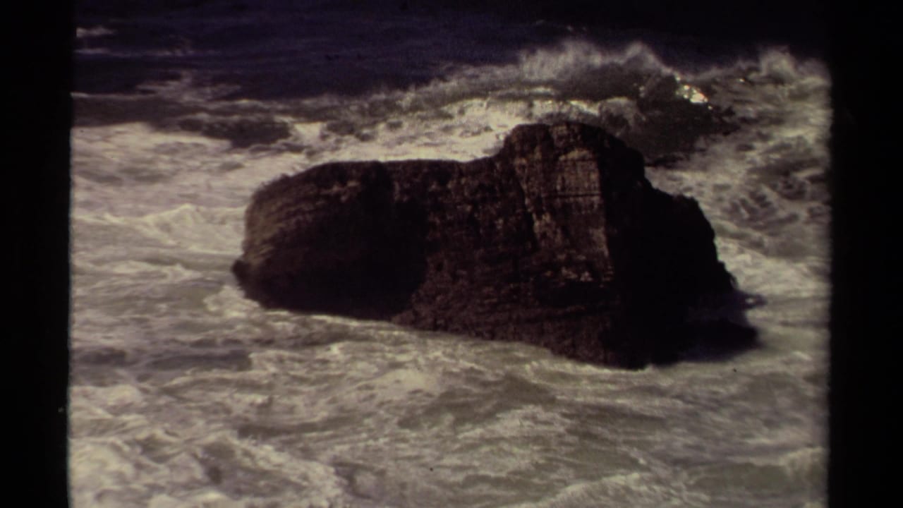 White surf curls around a large dark rock in rough water.