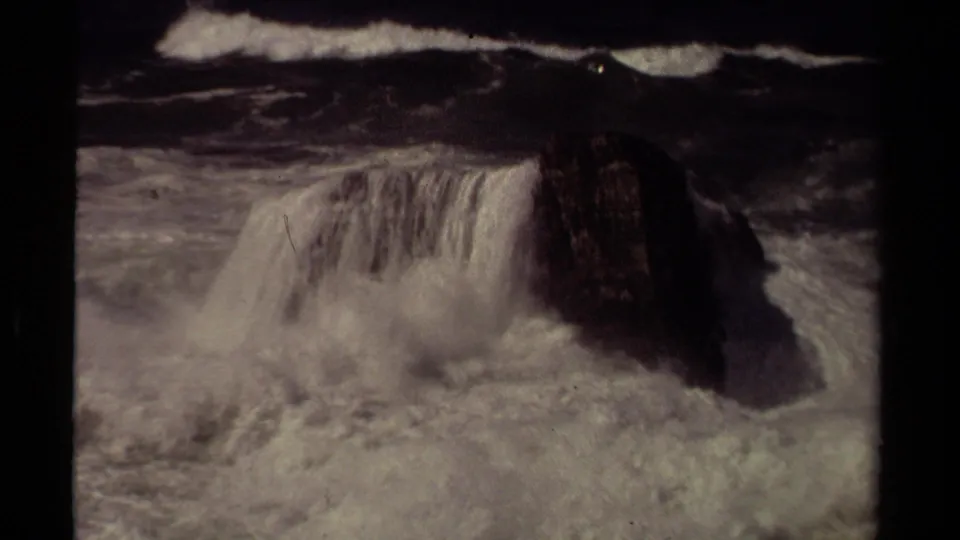 A dark coastal rock is partly covered by crashing white surf in 1980 8mm footage from Mendocino, California.