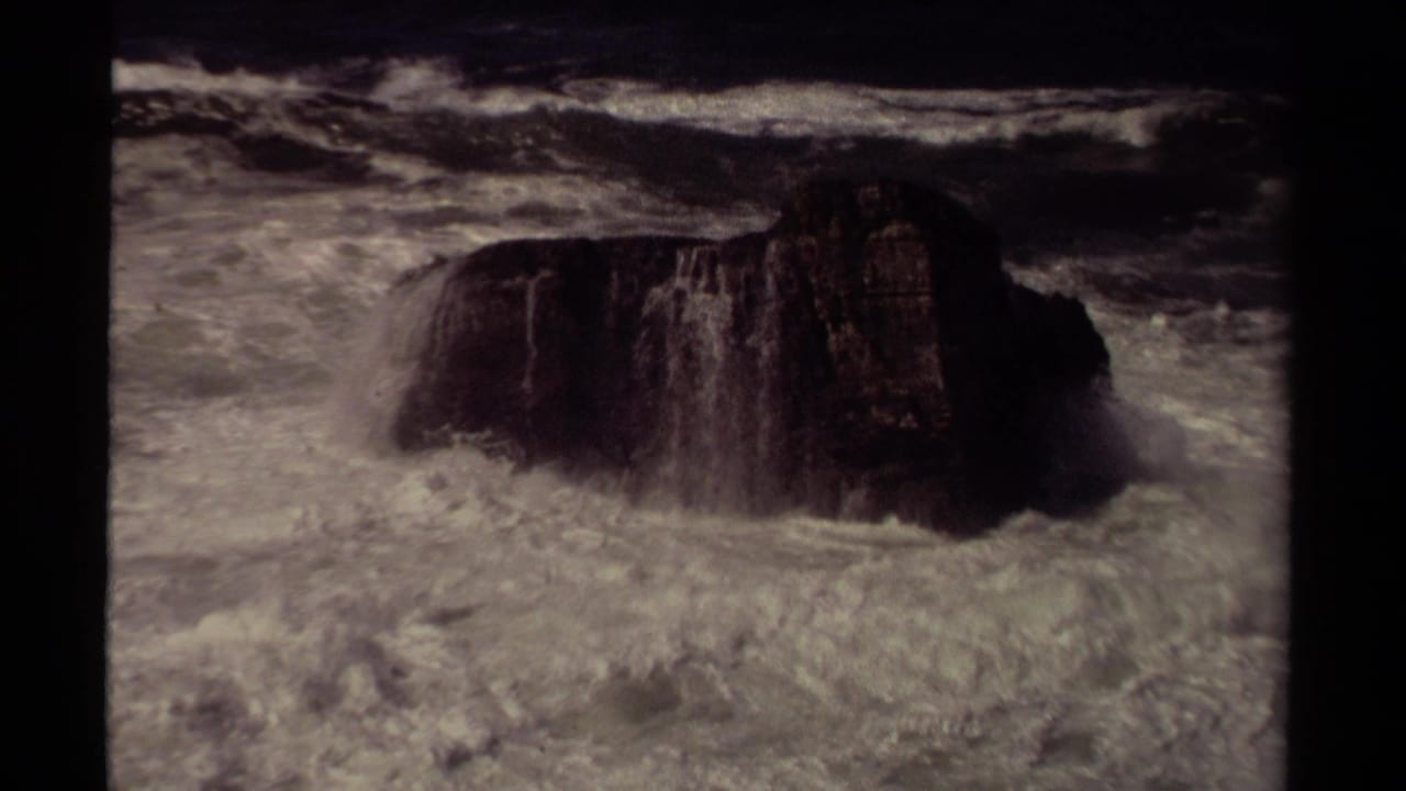 Mendocino-area 1980 8mm frame showing foamy surf rolling around a large dark coastal rock.