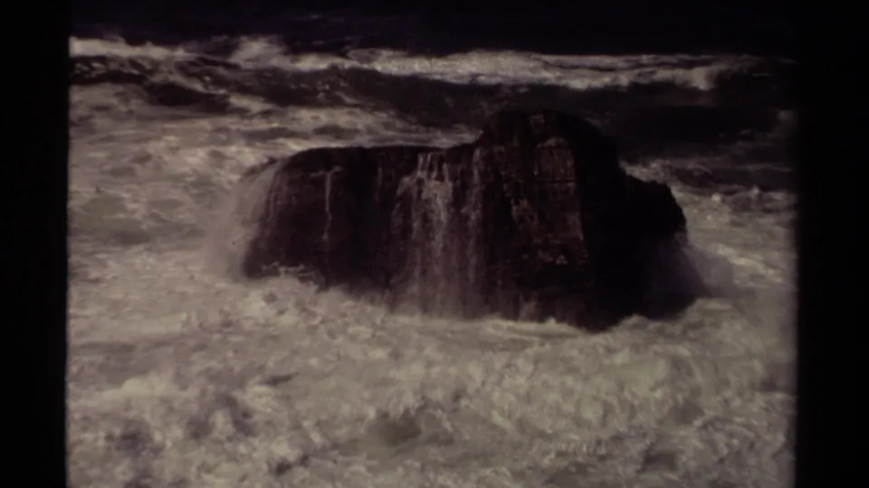 Mendocino-area 1980 8mm frame showing foamy surf rolling around a large dark coastal rock.