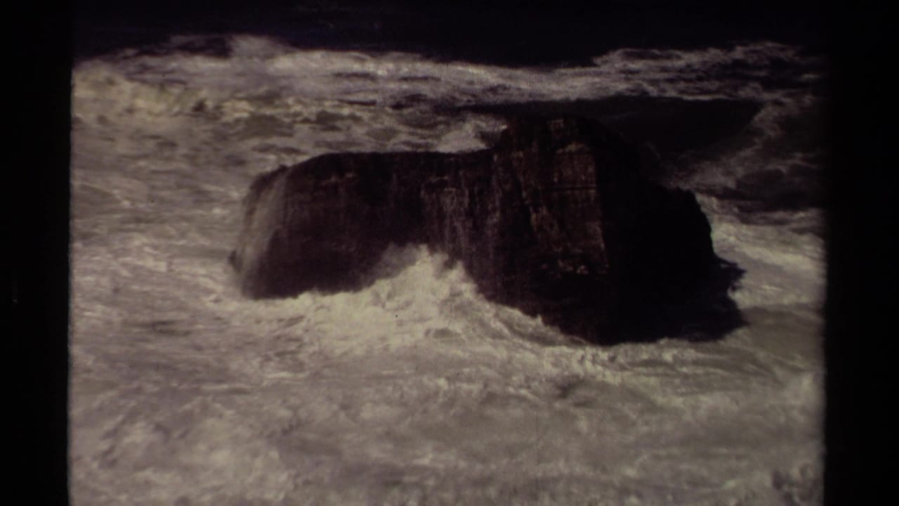 Archival 8mm view of rough Mendocino surf striking a dark rock formation in open water.