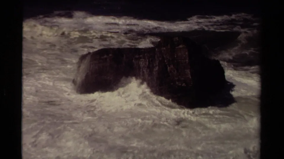 Archival 8mm view of rough Mendocino surf striking a dark rock formation in open water.